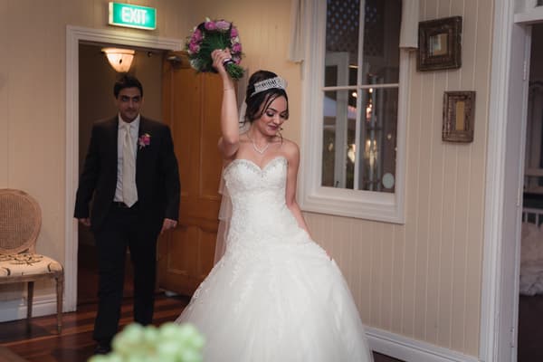 Maryam, the bride, raises her bouquet while walking indoors at Hillstone St Lucia — The Rosewood Room, followed by Pasha, the groom, in a suit with a boutonniere.