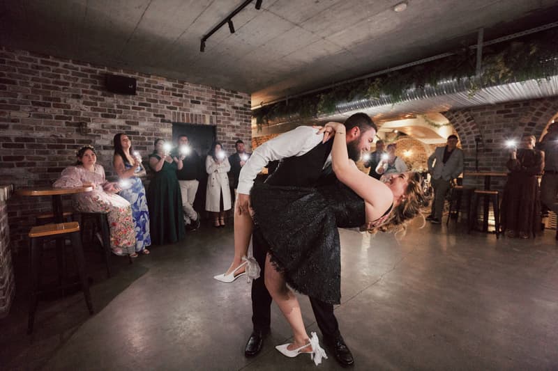 Christopher dips Jayme while dancing on the reception stage at Sandstone Point Hotel — The Cellar, with guests watching and taking photos.