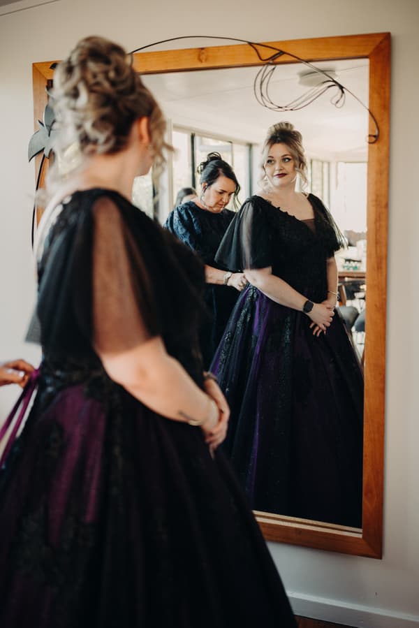 The bride stands in front of a large wooden-framed mirror at Ocean View Estates, wearing a dark purple and black gown with sheer sleeves, while another woman adjusts the back of her dress.