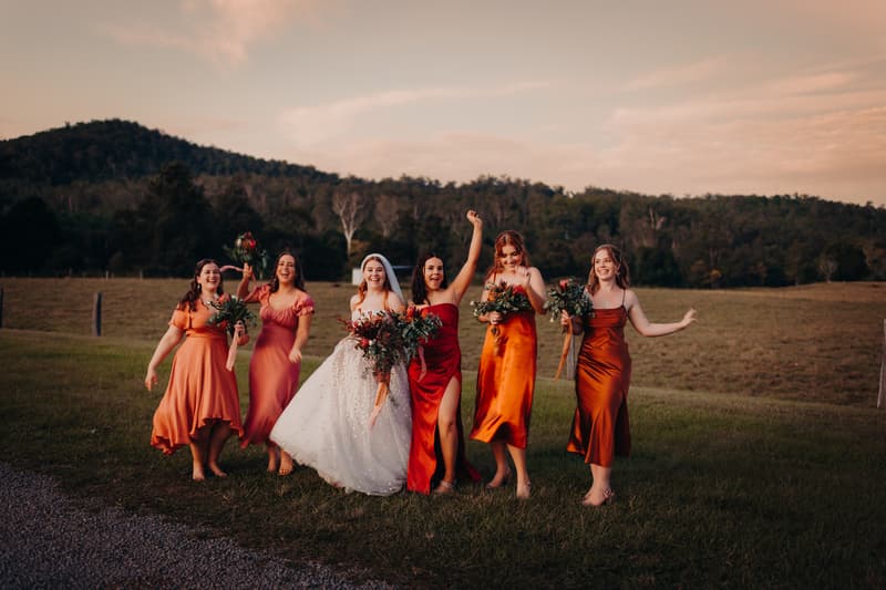 The bride Lilly and five bridesmaids in various shades of orange and red dresses walk together on grass at Yabbaloumba Retreat with a forested hill in the background during golden hour.