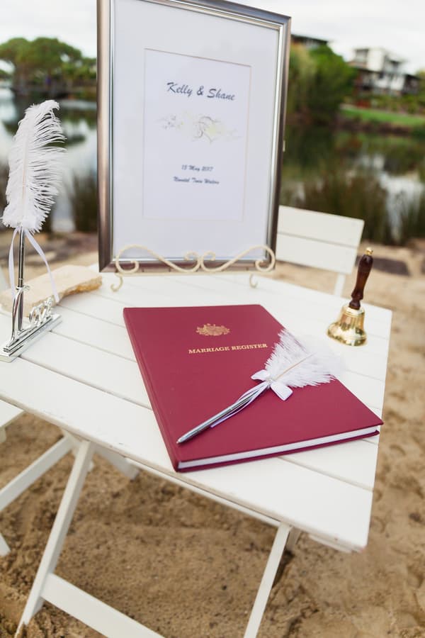 A white table at Novotel Sunshine Coast Resort holds a red marriage register book with a white feather pen on top, a framed sign with the names Kelly & Shane and the date 13 May 2017, and a small brass bell.