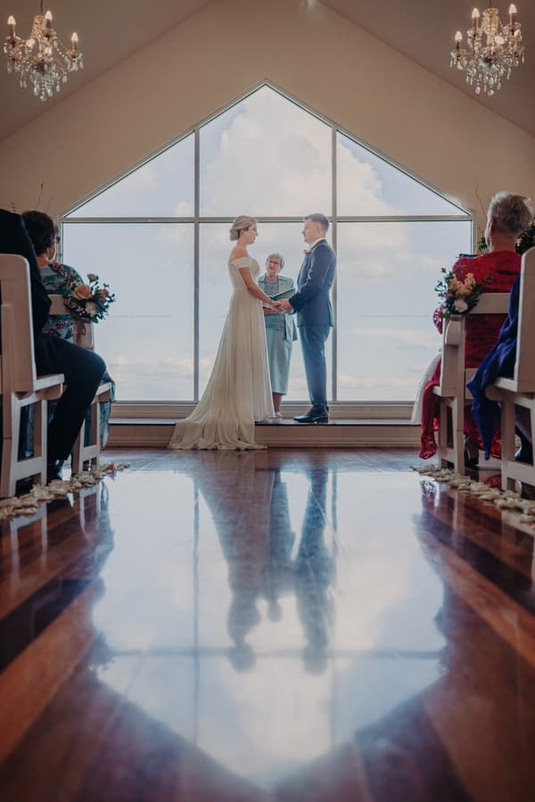 Bride Courtney and groom Liam hold hands facing each other at the altar with an officiant between them during their wedding ceremony at Tiffany's Maleny — Chapel, with guests seated on either side.