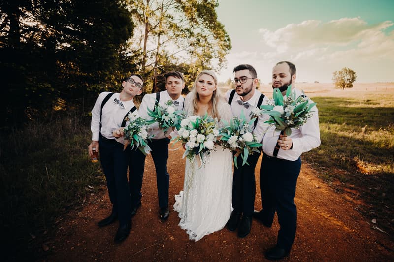 The bride Annie poses with four groomsmen outdoors on a dirt path at Ocean View Estates, each holding bouquets and making playful faces.