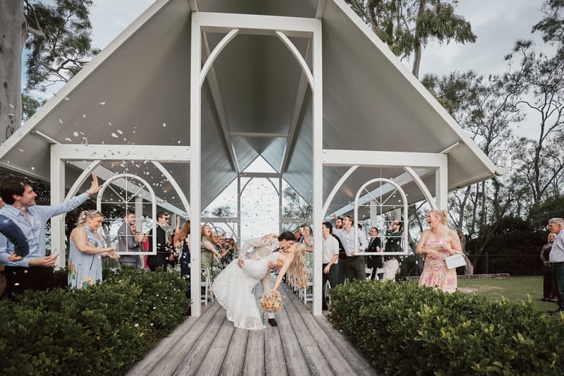 Morgan and Joel share a kiss at the Sandstone Point Hotel — The Pavilion ceremony stage as guests throw confetti and celebrate around them.
