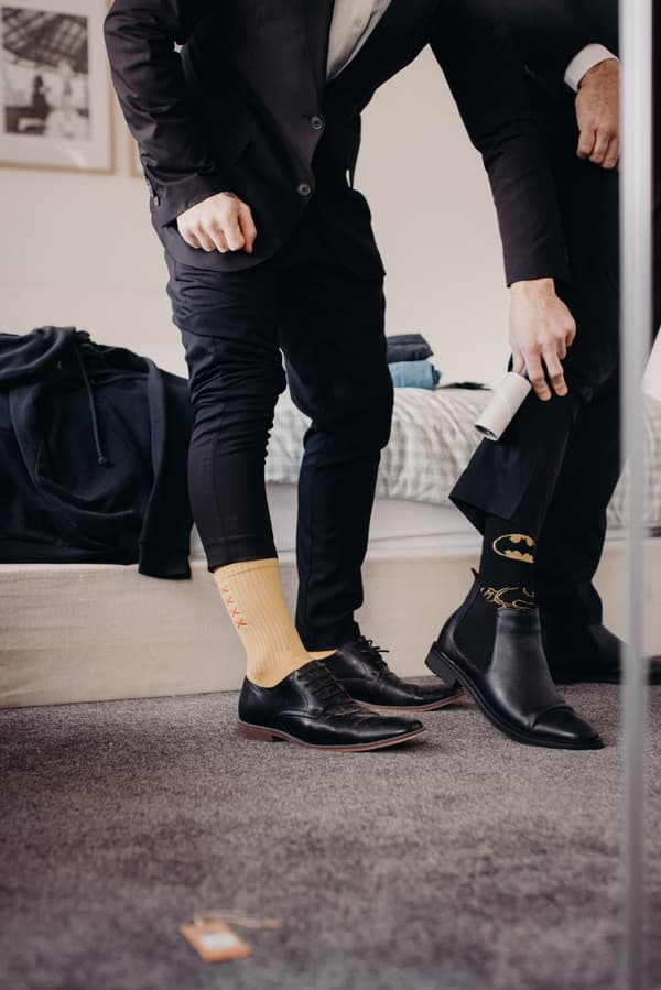 Two men in black suits stand indoors near a bed, one wearing yellow socks with red crosses and black dress shoes, the other wearing black Batman-themed socks and black dress shoes, using a lint roller on his pants.