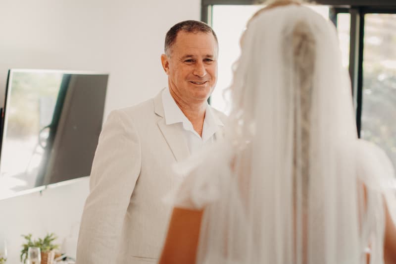 The bride's father smiles at the bride, who is seen from behind wearing a veil and wedding dress, inside a bright room at Sandstone Point Hotel.