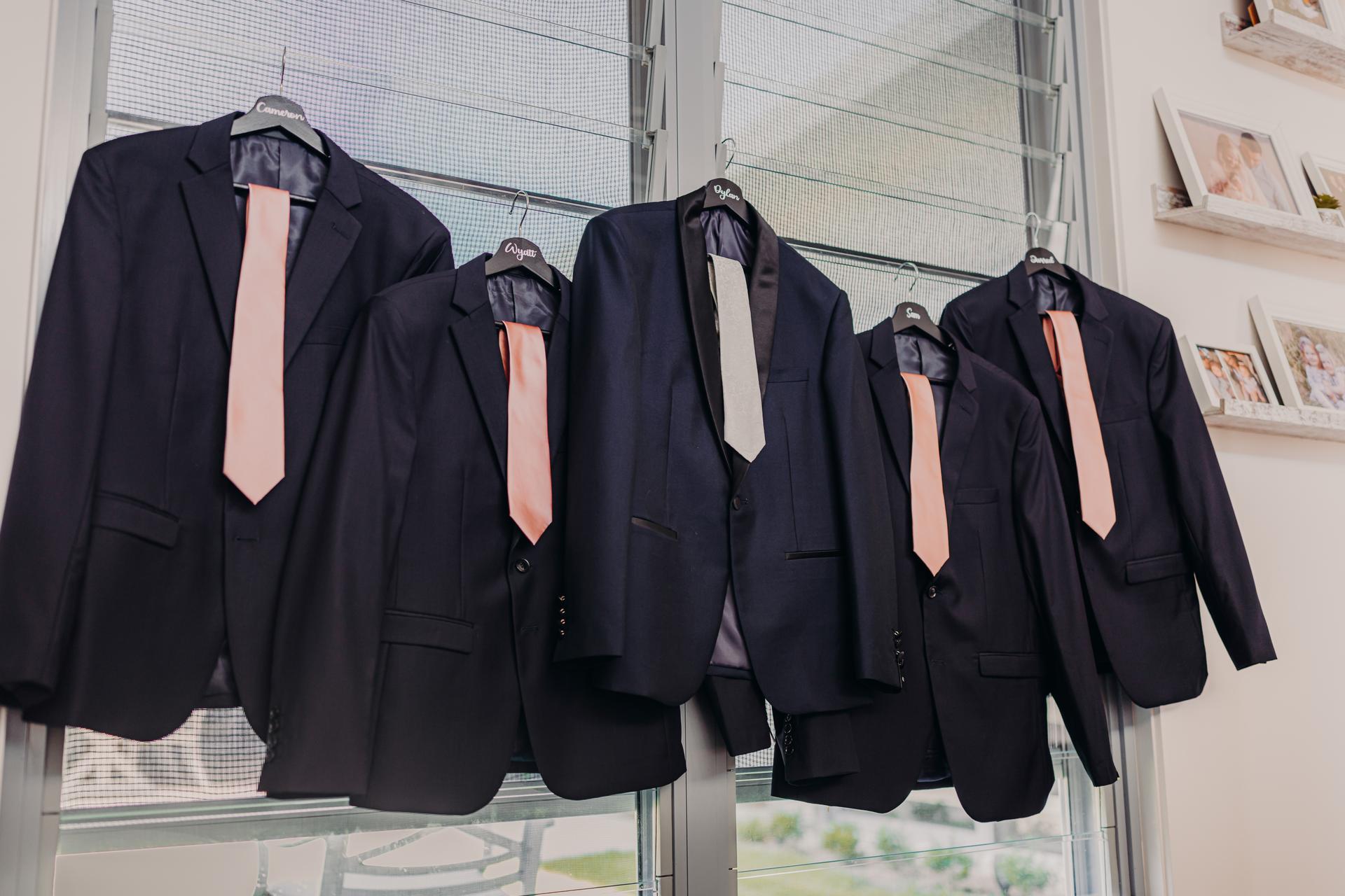 Five dark suits with ties hanging on hangers in front of a window, each hanger labeled with a name.