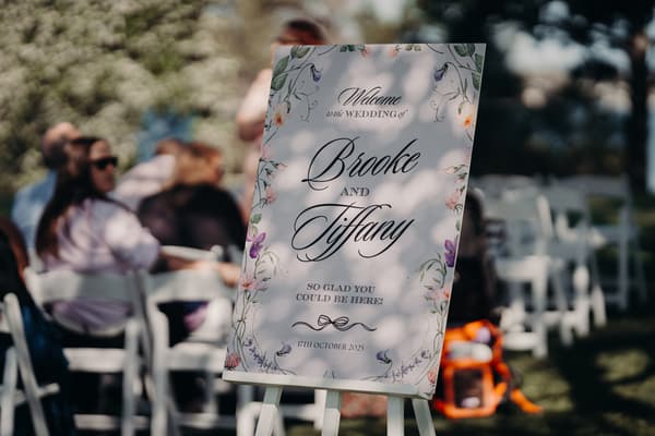 A floral wedding welcome sign for Brooke and Tiffany dated 17th October 2025 is displayed on an easel outdoors with seated guests in the background at Sandstone Point Hotel — The Pavilion.