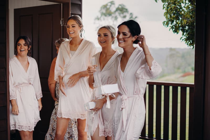 Four bridesmaids in matching light pink robes stand together near an open door at Preston Peak Winery Functions Centre, one holding a champagne flute and two holding small white boxes.