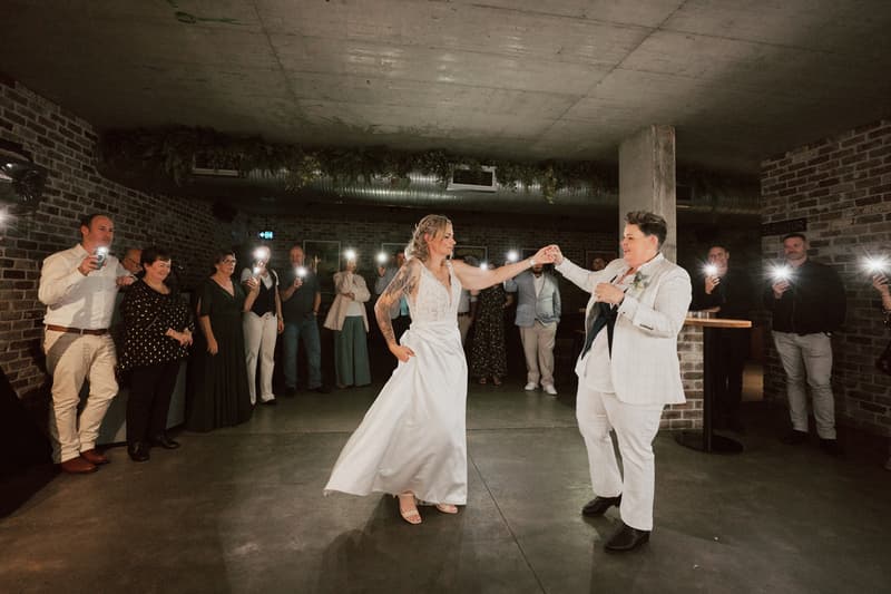 Brides Mandy and Selina dance together on the reception stage at Sandstone Point Hotel — The Cellar, surrounded by guests holding up phones with flashlights on.
