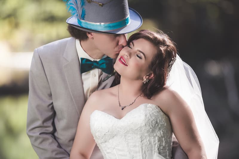 The groom Paul wearing a grey suit, teal bow tie, and grey hat with teal feather kisses the bride Holly on the cheek as she smiles with closed eyes in her white strapless wedding dress and veil at Kwila Lodge.