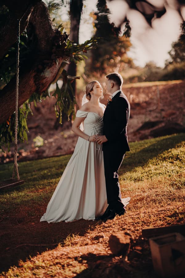 Courtney the bride and Liam the groom stand close together outdoors at Tiffany's Maleny during their couple portraits session, with Courtney holding a glass and a tree with a rope swing nearby.