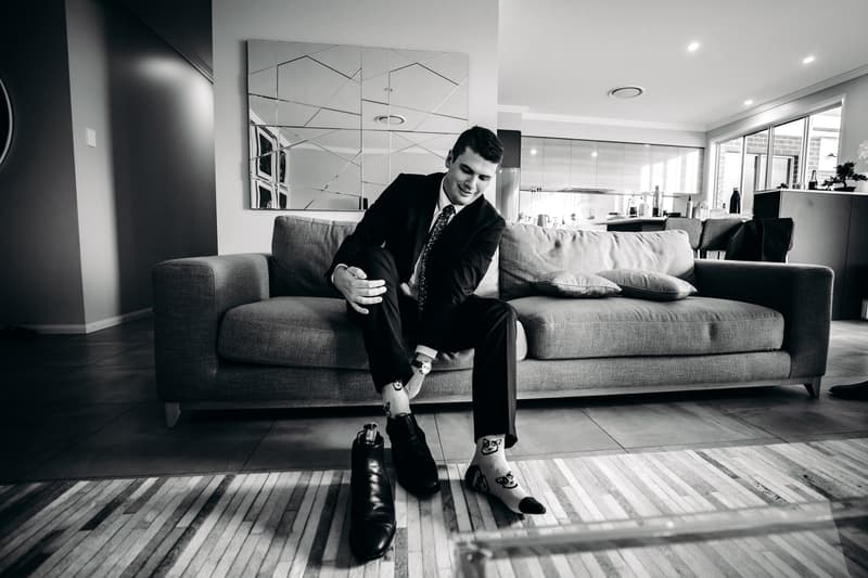 The groom Brent sits on a couch at Preston Peak Winery Functions Centre, putting on his black dress shoes and revealing patterned socks.