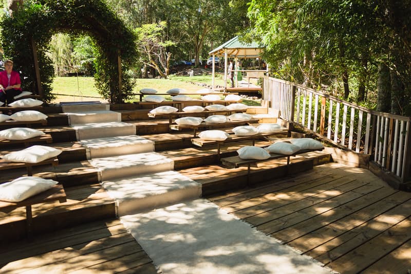 Empty outdoor ceremony seating area at Kwila Lodge with wooden benches, white cushions, and a white aisle runner under a leafy archway.