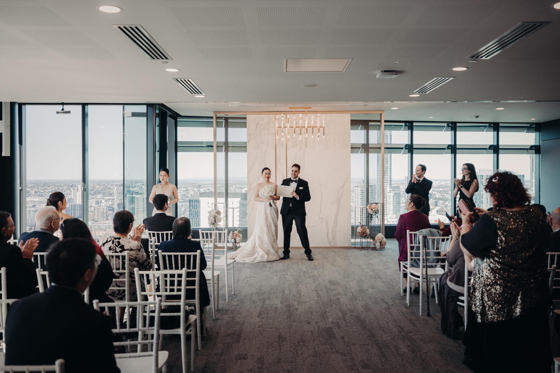The bride Aria and groom Antony stand at the ceremony stage at Brisbane registry ceremony room, holding a certificate while guests seated in rows applaud.