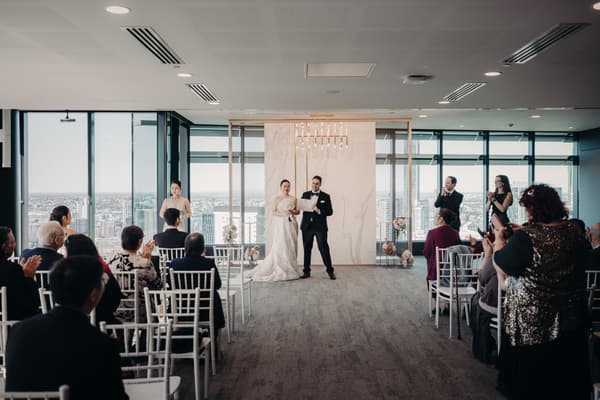 The bride Aria and groom Antony stand at the ceremony stage at Brisbane registry ceremony room, holding a certificate while guests seated in rows applaud.
