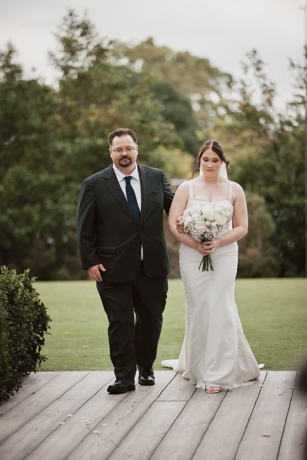The bride Terion, holding a bouquet of white flowers, walks arm-in-arm with an older man on a wooden pathway at Sandstone Point Hotel — The Pavilion, with greenery in the background.