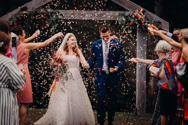 Bride Lilly and groom Connor walk hand in hand under a wooden arch decorated with flowers at Yabbaloumba Retreat — By The River as guests throw confetti.