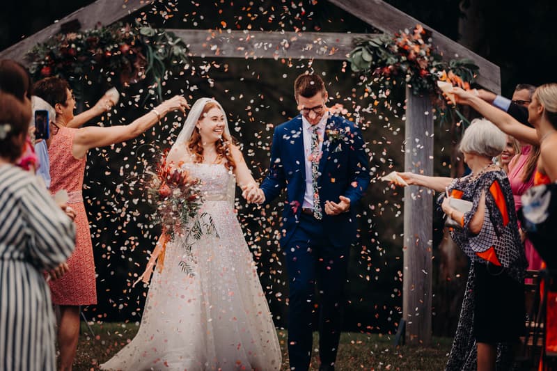 Bride Lilly and groom Connor walk hand in hand under a wooden arch decorated with flowers at Yabbaloumba Retreat — By The River as guests throw confetti.