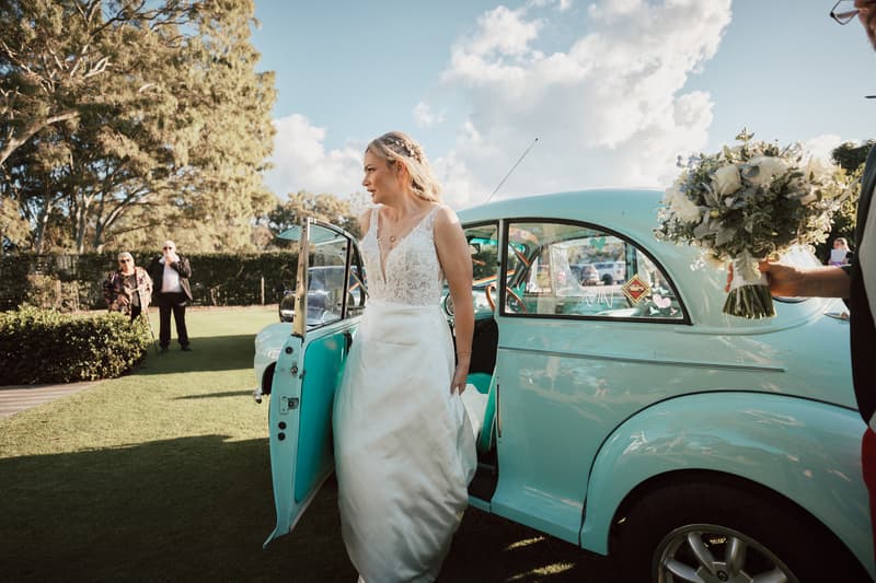 The bride Mandy steps out of a vintage light blue car on the lawn at Sandstone Point Hotel — The Pavilion, holding her wedding dress while a person holds a bouquet nearby. Two guests stand in the background.