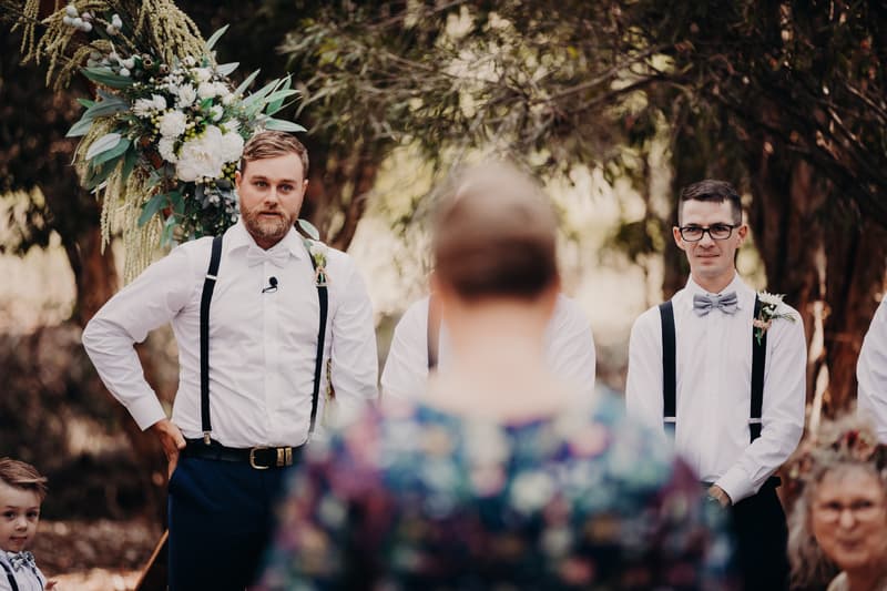 The groom Trent and a groomsman stand at the ceremony stage at Ocean View Estates, with a floral arrangement behind them and a blurred guest in the foreground.