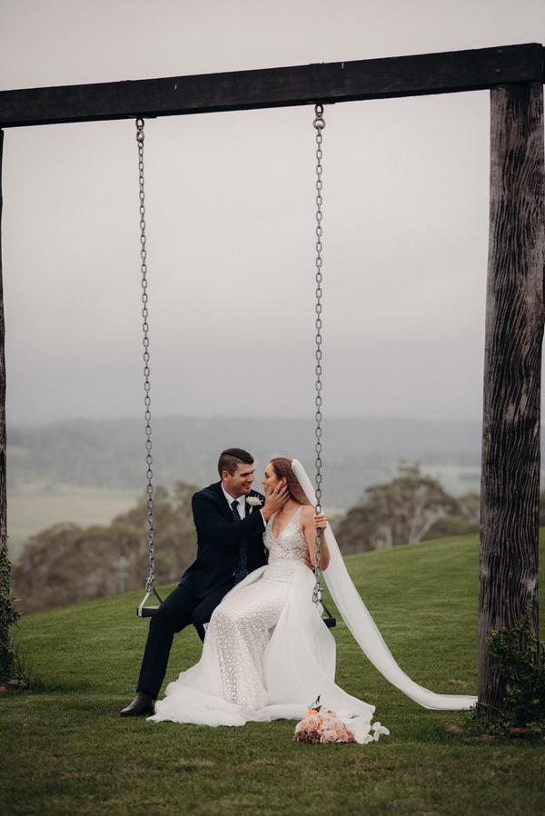 Bride Chelsea and groom Brent sit together on a wooden swing outdoors at Preston Peak Winery Functions Centre, with a scenic background of trees and hills. Chelsea wears a white wedding dress with a long veil and holds the swing chain, while Brent in a dark suit gently touches her face. A bridal bouquet lies on the grass nearby.