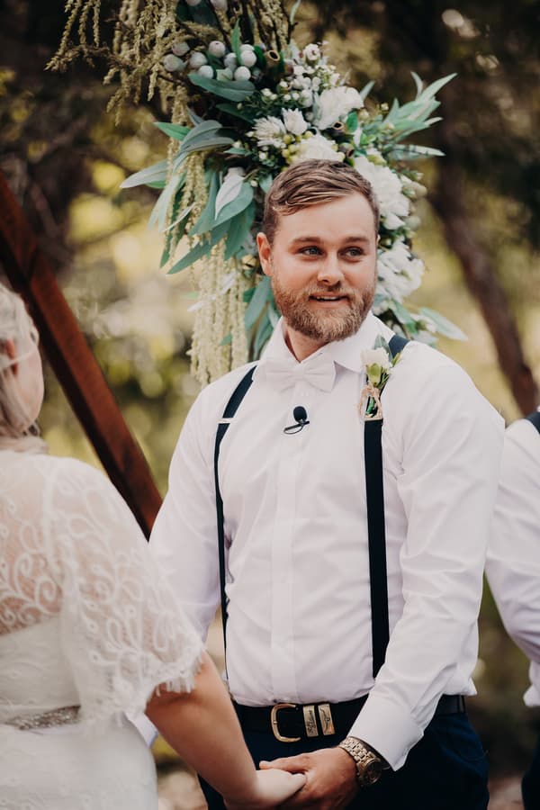 Trent, the groom, holds hands with Annie, the bride, during their wedding ceremony at Ocean View Estates. Trent is wearing a white shirt with black suspenders and a boutonniere, standing in front of a floral arrangement.