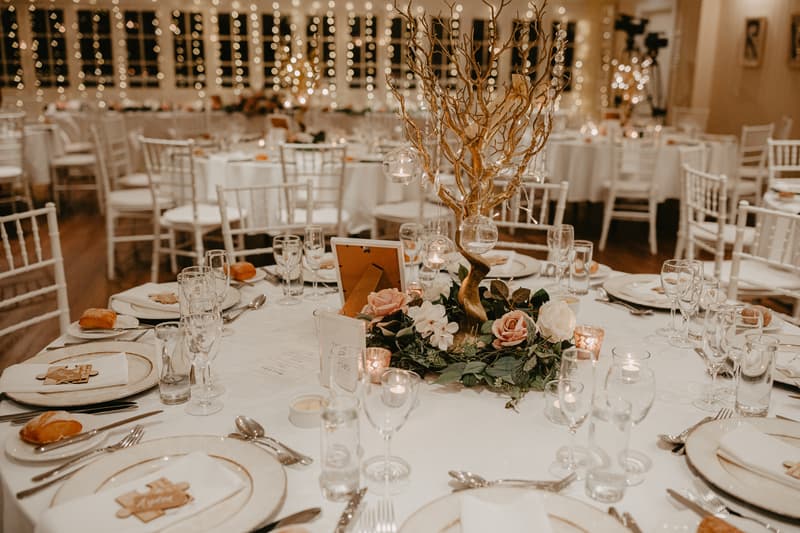 Round tables set for a wedding reception with white tablecloths, floral centerpieces featuring a gold decorative tree, candles, glassware, plates, cutlery, and bread rolls on napkins.