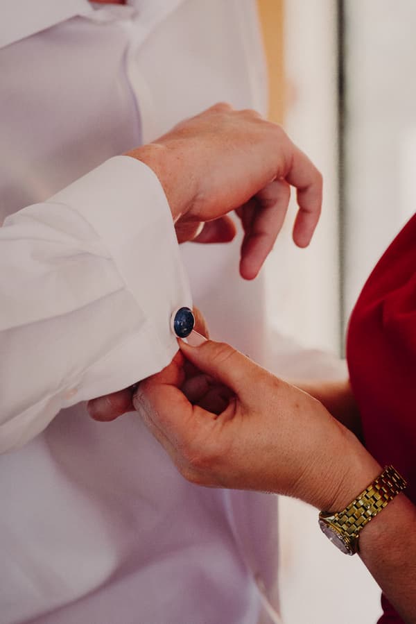 A person fastens a cufflink on the groom Arran's white shirt sleeve during preparation.