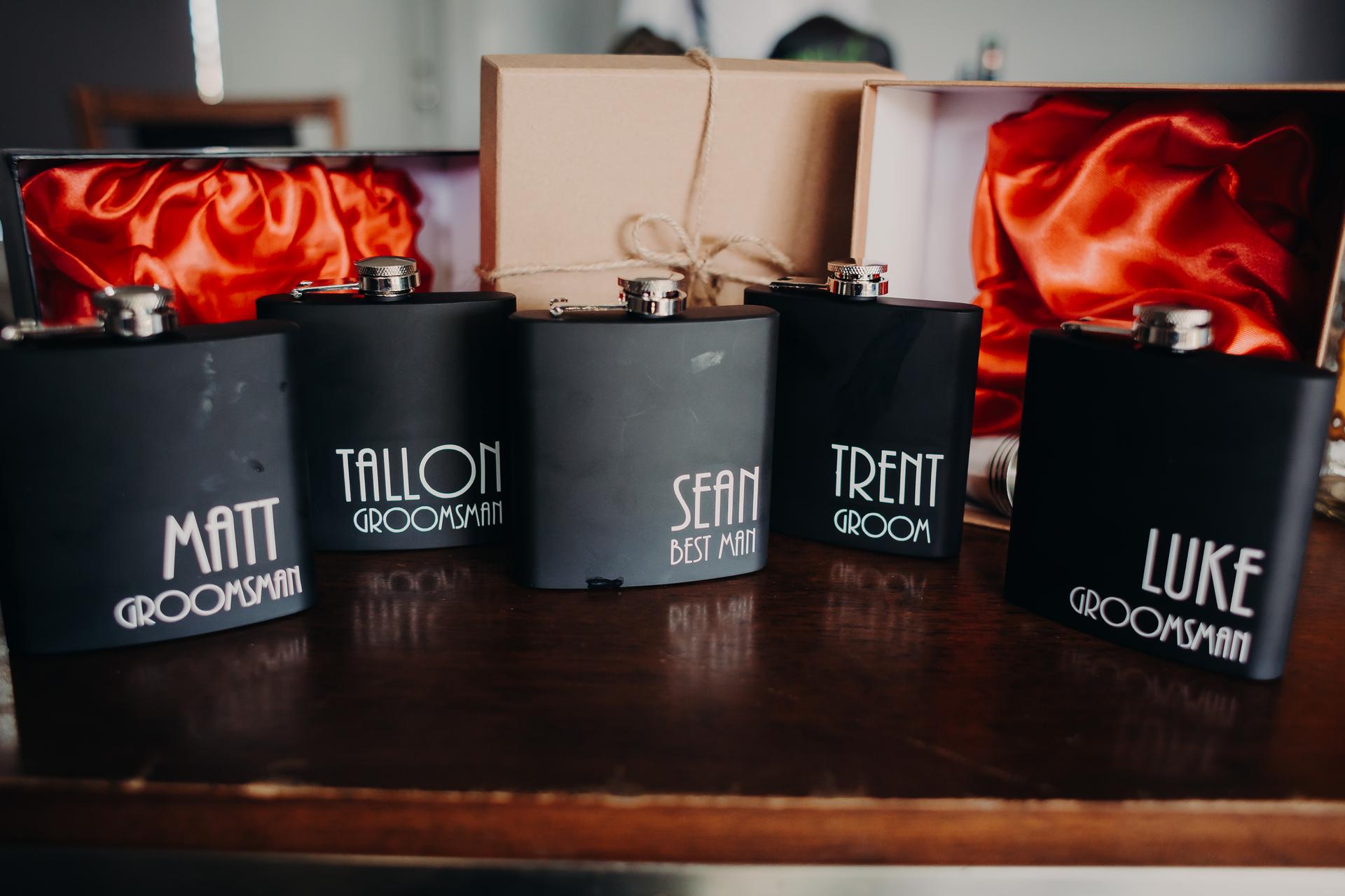 Five black flasks labeled with names and roles: Matt (Groomsman), Tallon (Groomsman), Sean (Best Man), Trent (Groom), and Luke (Groomsman) displayed on a wooden surface with gift boxes lined with red satin fabric in the background at Ocean View Estates.