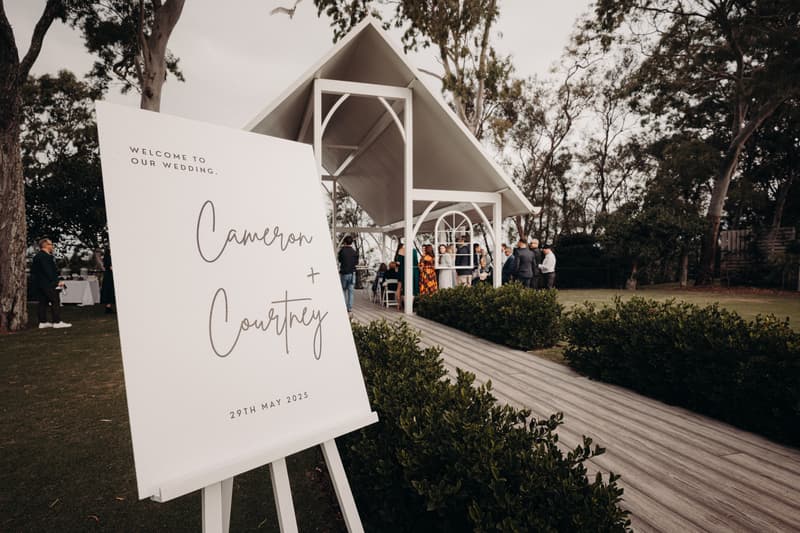Wedding welcome sign for Cameron and Courtney dated 29th May 2025 is displayed on an easel outside the pavilion at Sandstone Point Hotel, with guests gathered inside and around the pavilion.