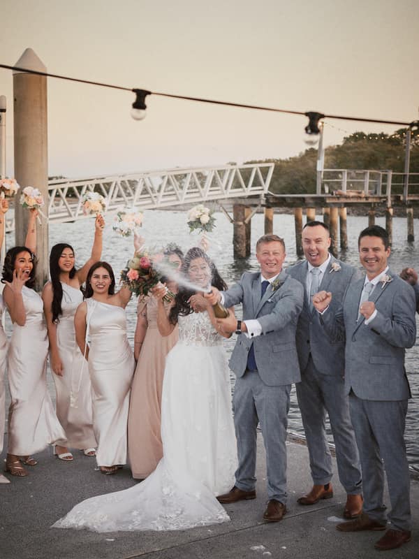 The bride Jheng and groom Dane celebrate with their bridal party at Sandstone Point Hotel, The Pavilion, as Dane sprays champagne while bridesmaids hold bouquets and groomsmen cheer.