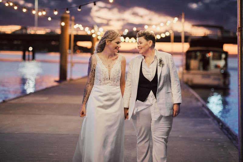 Mandy and Selina walk hand in hand along a pier at sunset with string lights overhead and water on both sides.