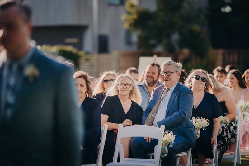 Guests seated outdoors at Sandstone Point Hotel — Rustic Arbour during the wedding ceremony, with a blurred figure of the groom in the foreground.