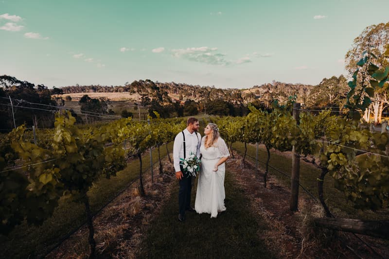 Annie and Trent stand together between rows of grapevines at Ocean View Estates, with Annie holding a bouquet and both looking at each other.