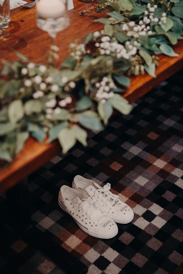 A pair of white sneakers with decorative studs and text placed on a checkered carpet floor beneath a wooden table adorned with green foliage and white berries at Sandstone Point Hotel.