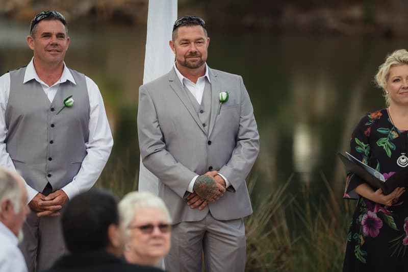The groom Shane stands at the ceremony stage at Novotel Sunshine Coast Resort wearing a light grey suit with a white shirt and boutonniere, accompanied by a groomsman in a grey vest and white shirt, and a female celebrant holding a book.