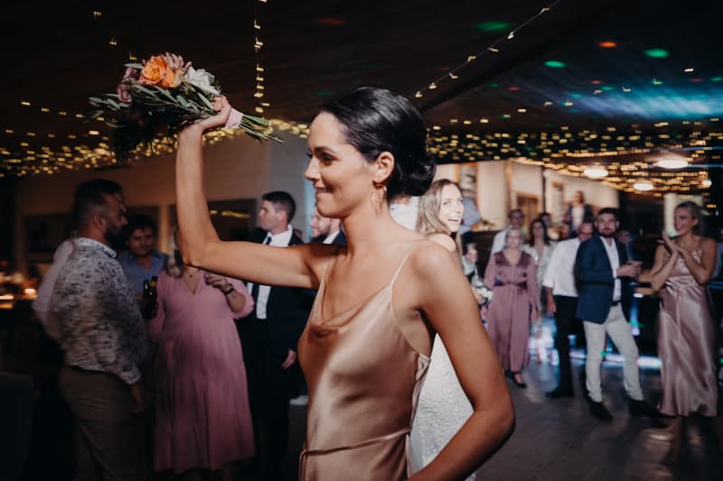 A bridesmaid in a satin dress holds a bouquet aloft at Preston Peak Winery Functions Centre reception with guests in the background.