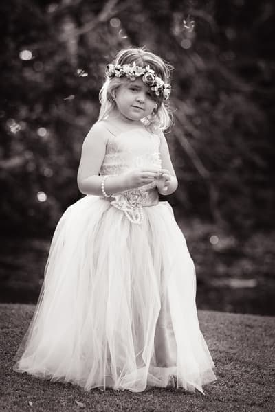 A young flower girl wearing a floral crown and a long tulle dress stands outdoors on grass. Wedding family photos by She Said Yes.