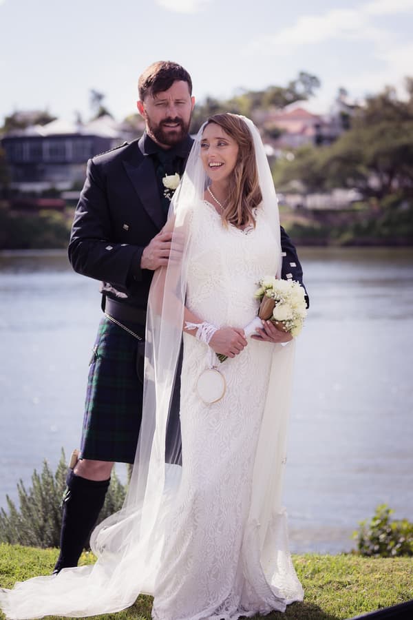 Francesca the bride in a white lace gown and veil holding a bouquet stands in front of Ben the groom wearing a black jacket and tartan kilt by a riverside outdoors.