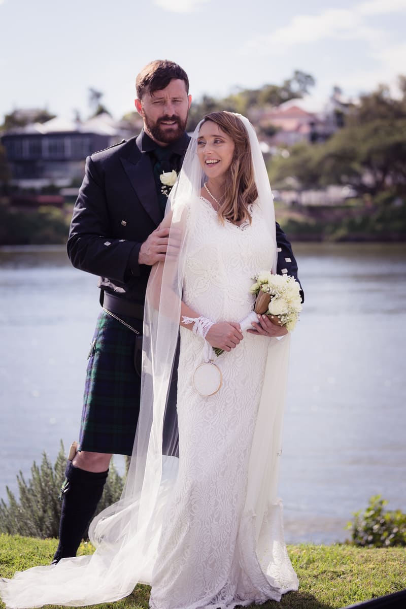 Francesca the bride in a white lace gown and veil holding a bouquet stands in front of Ben the groom wearing a black jacket and tartan kilt by a riverside outdoors. Wedding photographer and videographer by She Said Yes.