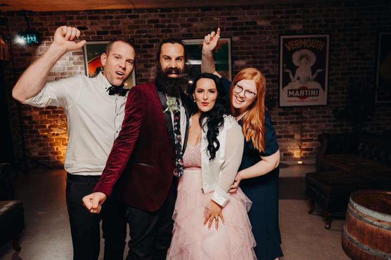 David in a burgundy jacket and floral tie stands next to Mindy in a pink dress and white jacket, accompanied by two guests, one in a white shirt and bow tie and the other in a navy dress, posing together at the Sandstone Point Hotel — Cellar during the reception.