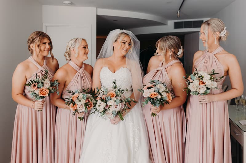 Emily the bride stands indoors at Sandstone Point Hotel Pavilion with four bridesmaids in matching blush pink dresses, all holding bouquets of flowers.