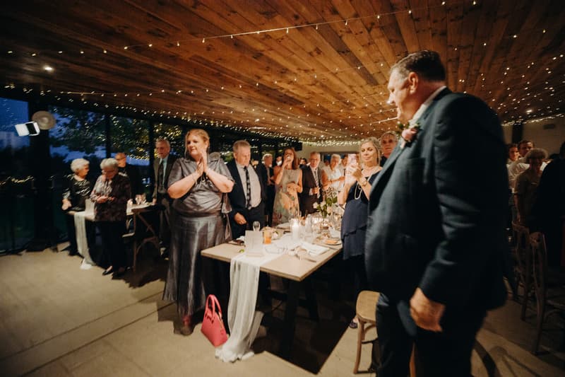 Guests stand and applaud at the Preston Peak Winery Functions Centre reception, with a man in a suit in the foreground and tables set with candles and floral arrangements.