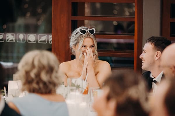 The bride Libby wearing heart-shaped sunglasses on her head sits at a reception table at The Tides — The Pandanus Room, smiling and covering her mouth with her hands, while the groom Kyle in a tuxedo smiles at her across the table.