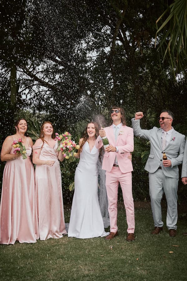 Olivia in her wedding dress and Jake in a pink suit celebrate with bridesmaids in pink dresses and a groomsman in a light grey suit as Jake pops a bottle of champagne outdoors at Eatons Hill Hotel — Lakeside.
