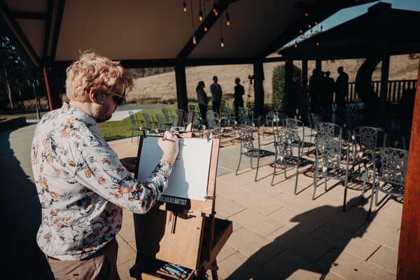 An artist sketches on an easel under a pavilion at Ocean View Estates — On The Lake, with empty chairs arranged for a ceremony and a few guests standing in the background.