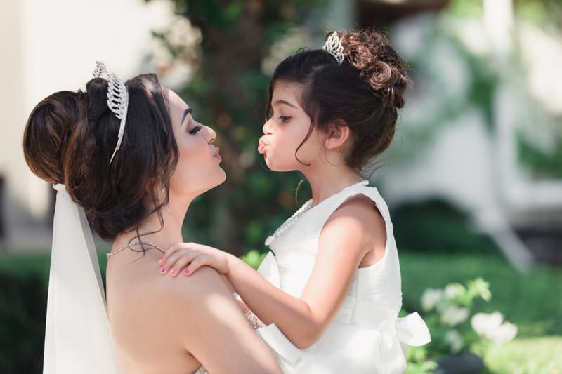 The bride at Hillstone St Lucia — The Quartyard holds a young girl dressed in white, both wearing tiaras and puckering their lips toward each other.