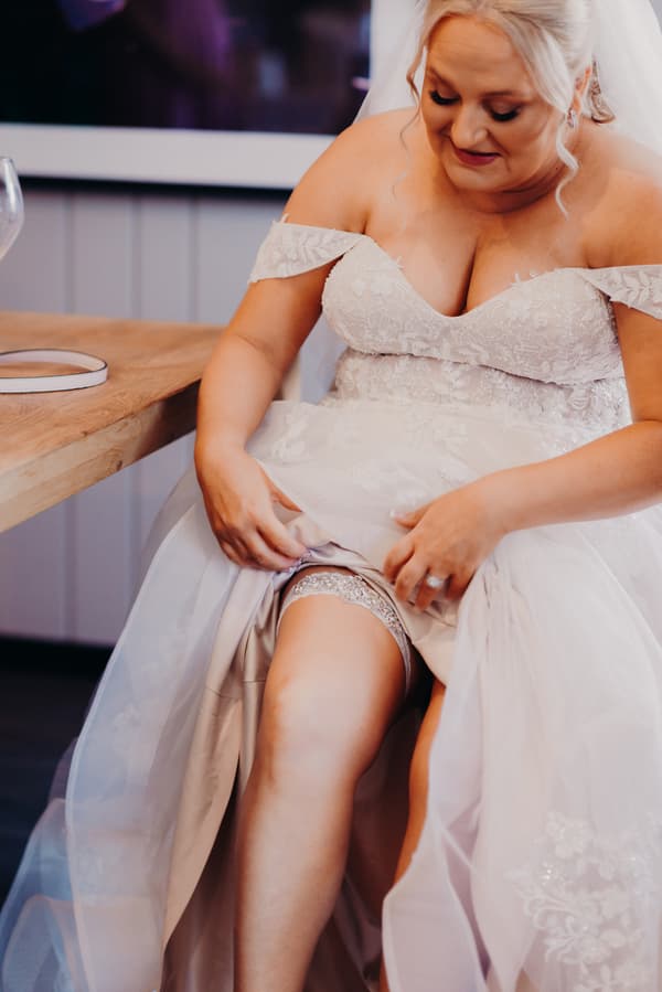 The bride Chantelle sits indoors at Sandstone Point Hotel, adjusting her lace garter on her leg while wearing an off-shoulder wedding gown.