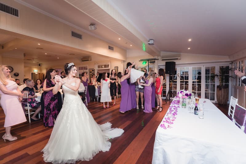 The bride in a white gown stands smiling in the center of Hillstone St Lucia — The Rosewood Room with guests and bridesmaids in purple dresses gathered behind her, some holding a flower girl lifted in the air near the dance floor.
